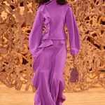 Model in purple outfit walks runway with handbag.