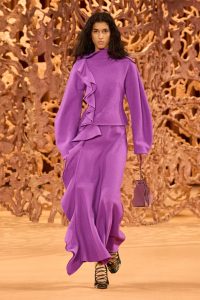 Model in purple outfit walks runway with handbag.