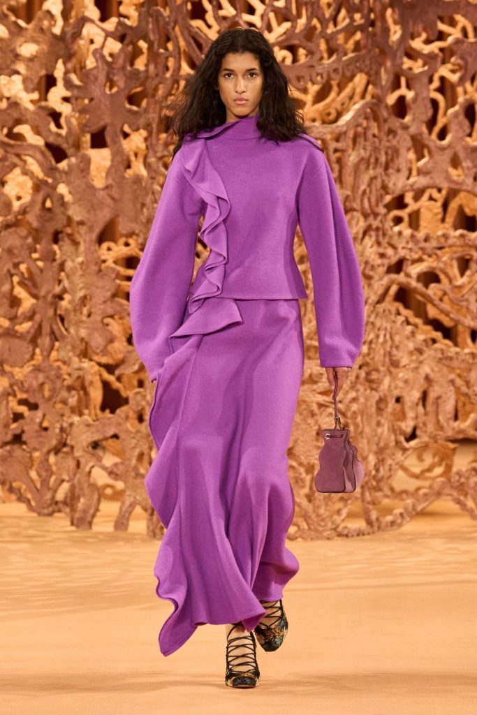 Model in purple outfit walks runway with handbag.